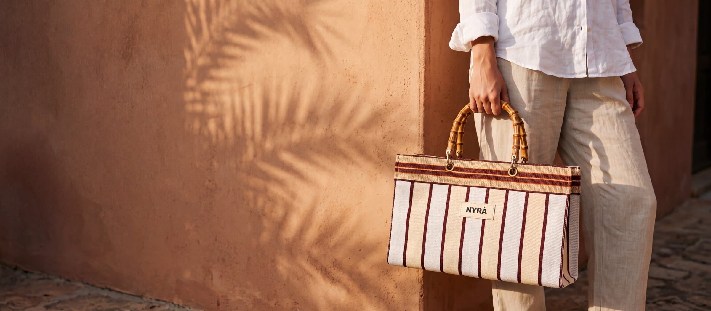 Striped recycled nylon tote with a bamboo handle, carried against a sunlit terracotta wall